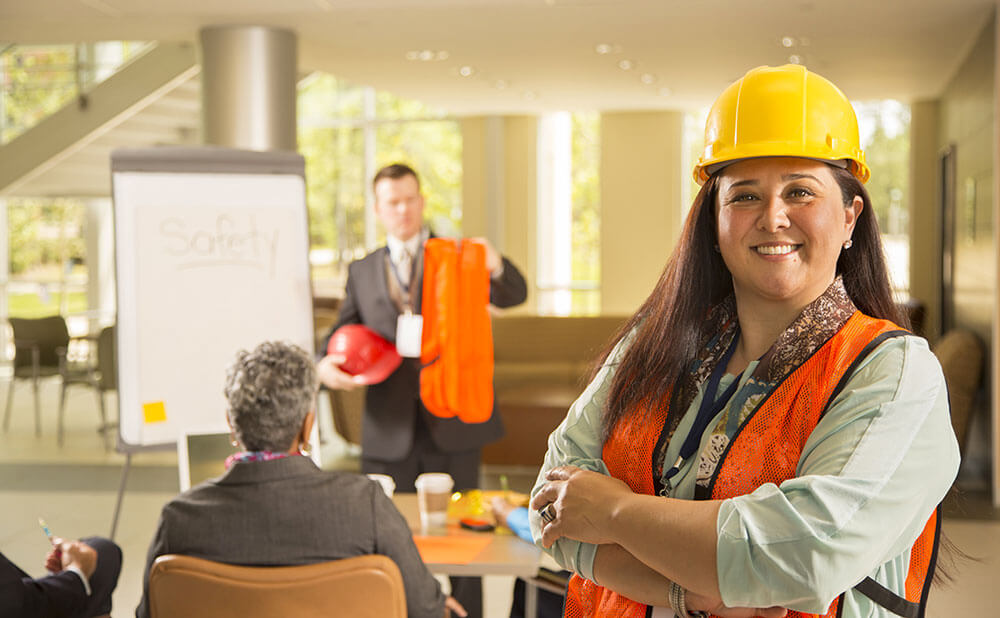Formation professionnelle Une femme en tenue de travail de sécurité sourit. Derrière elle il y a une réunion et un homme présente du matériel de sécurité.